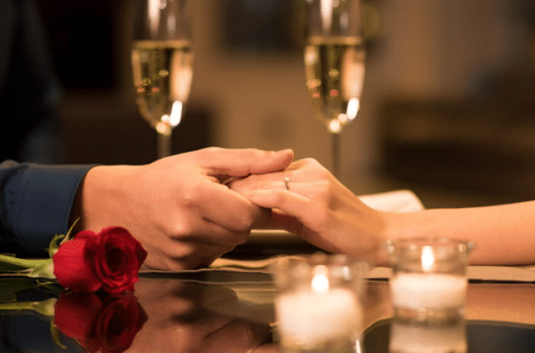 How to Plan a Romantic Vacation in Las Vegas 1 A closeup of a couple holding hands on a restaurant table with two glasses of champagne in background. There are also candles and a rose on the table.