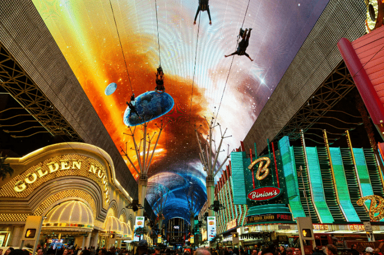 Top 13 Hidden Las Vegas Gems 1 a lighted canopy photo of the Fremont Street Experience as zipliners go by. Beneath the ziplines is a large crowd standing outside the Golden Nugget which is a hotel and casino.